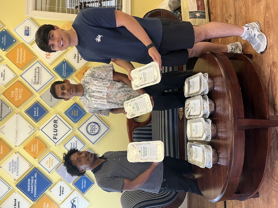 Sid Sharma and two volunteers holding labeled Waste2Taste banana bread loaves in a community center lobby